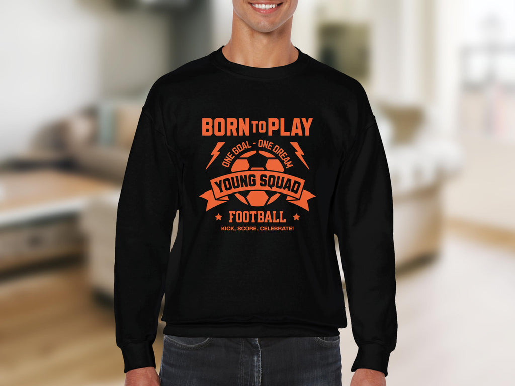 In a cozy room, someone wears Garment Graphics black football sweatshirt with orange text: Born to Play, One Goal - One Dream, Young Squad, Football, Kick, Score, Celebrate. Its ideal for young aspiring footballers and comes in unisex sizes.