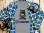 A gray Garment Graphics t-shirt with a Scania truck and logo lies on a wooden surface, ideal for truck fans. Its paired with a blue plaid shirt, headphones, camera, and coffee cup. This graphic crewneck sweatshirt is perfect for a casual day out.