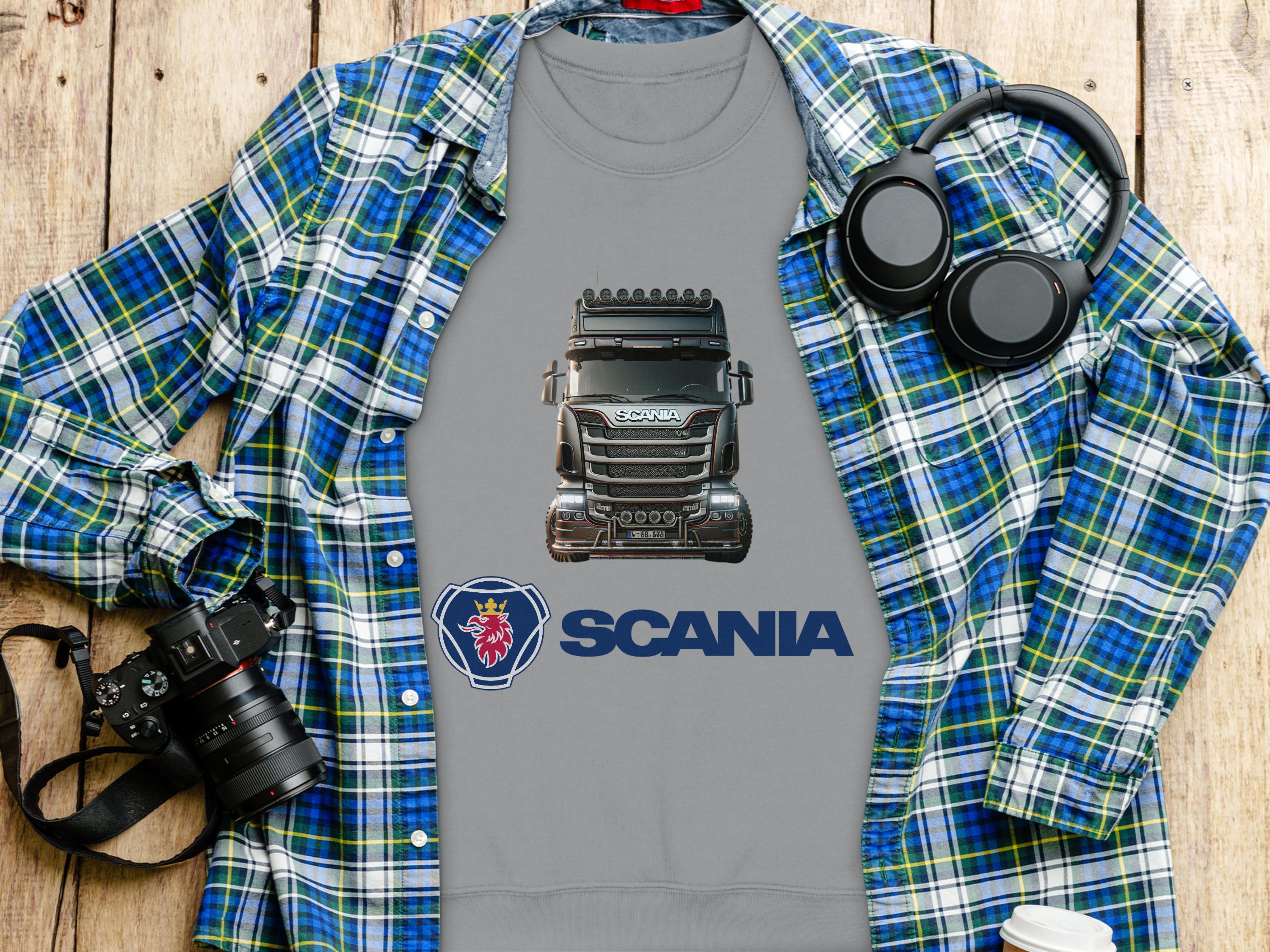 A gray Garment Graphics t-shirt with a Scania truck and logo lies on a wooden surface, ideal for truck fans. Its paired with a blue plaid shirt, headphones, camera, and coffee cup. This graphic crewneck sweatshirt is perfect for a casual day out.