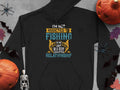 A black fishing hoodie by Garment Graphics with the text Im not addicted to fishing, were just in a very committed relationship in various fonts and colors, featuring pumpkins, bats, a skeleton, and autumn leaves.