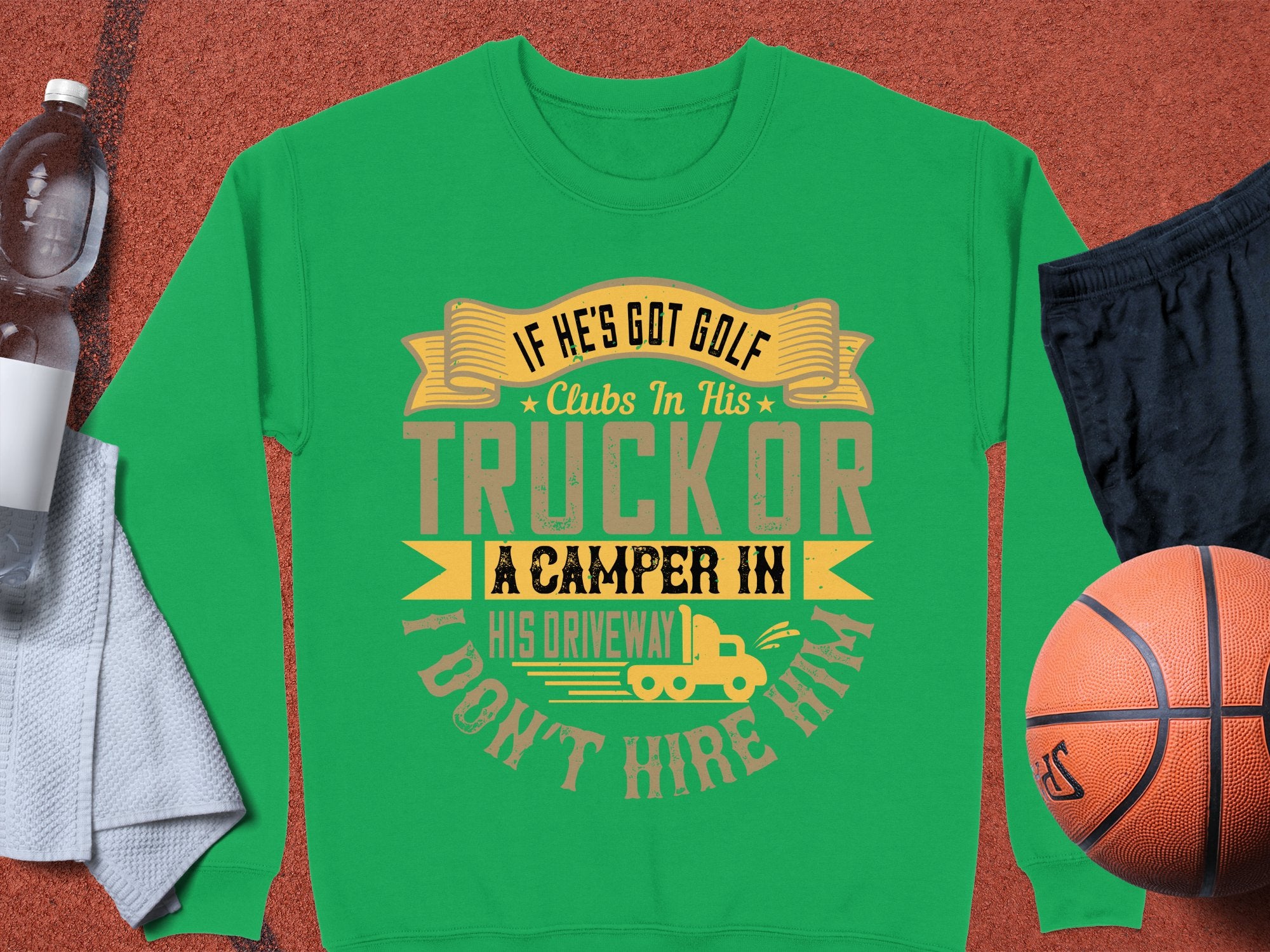A green Garment Graphics sweatshirt features the text, If hes got golf clubs in his truck or a camper in his driveway, I dont hire him. Its paired with black shorts, a basketball, a water bottle, and a grey towel—ideal for those who enjoy mixing outdoor golf with camping adventures.