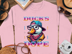 A bold pink shirt from Garment Graphics features a duck in glasses, a cap, and scarf with Ducks Dope surrounded by Brave, Cute, Smart, and Cool. Ideal for adventurous days, it rests on a wooden surface alongside travel items.