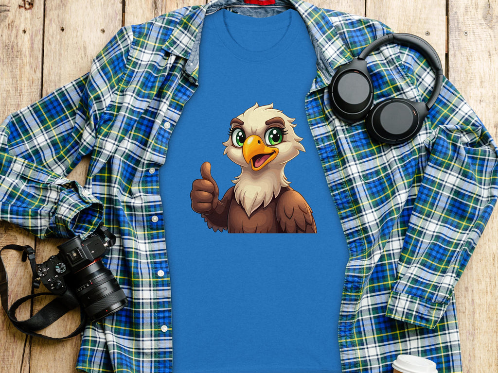 A Garment Graphics Eagle T-shirt with a cartoon bald eagle giving a thumbs-up lies on a wooden surface, surrounded by casual staples: a blue and green plaid shirt, black headphones, a camera, and a white takeaway coffee cup.