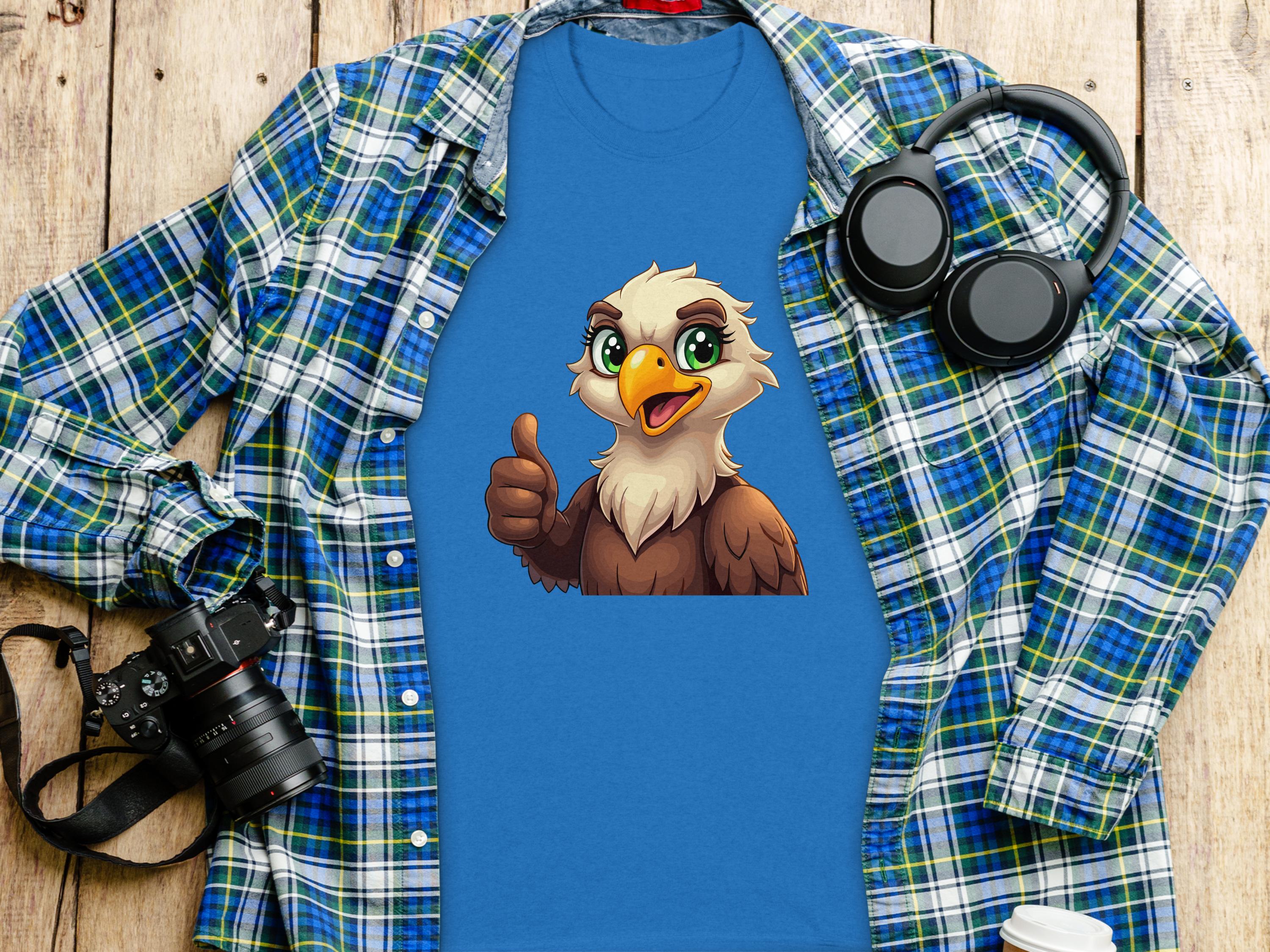 A Garment Graphics Eagle T-shirt with a cartoon bald eagle giving a thumbs-up lies on a wooden surface, surrounded by casual staples: a blue and green plaid shirt, black headphones, a camera, and a white takeaway coffee cup.