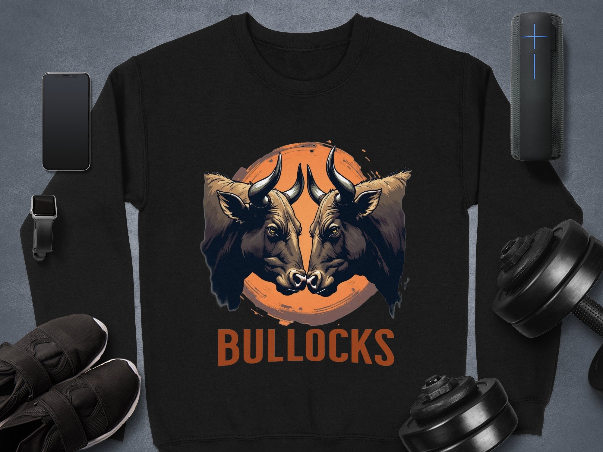 The Garment Graphics black sweatshirt features a striking bullock design with two bulls facing each other and an orange sun-like circle above the word BULLOCKS. Unisex sizing makes it versatile, pairing well with a smartphone, smartwatch, shoes, speaker, or dumbbells for any style.