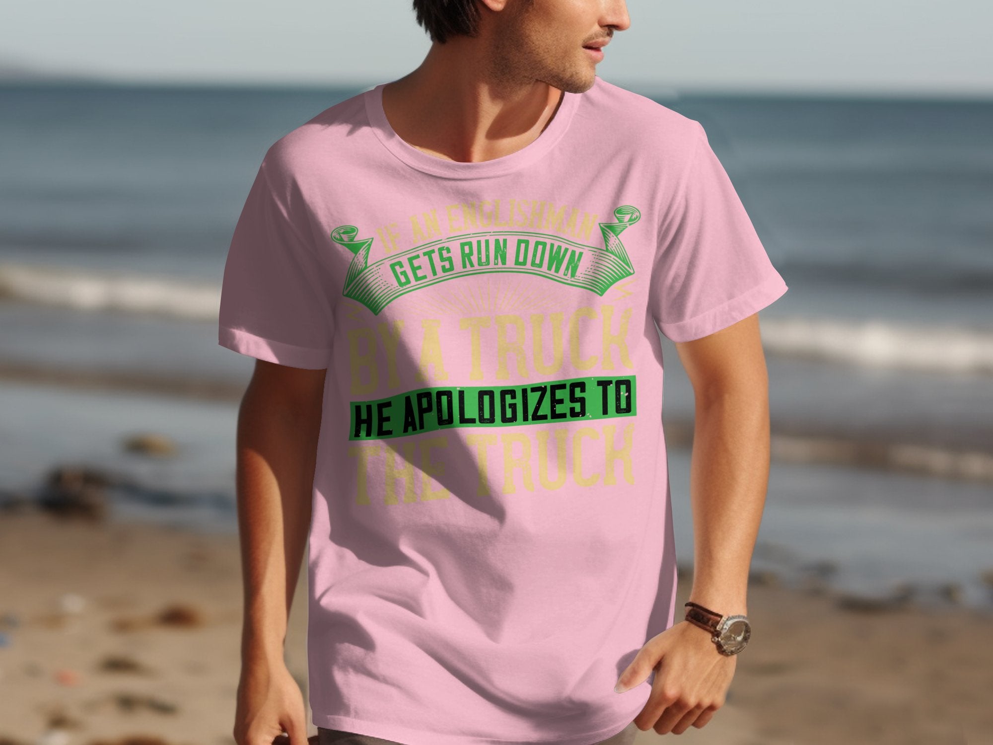 Someone is wearing a Garment Graphics pink T-shirt that humorously says, If an Englishman gets run down by a truck, he apologizes to the truck. They are standing on a beach with sand and water visible, humorously highlighting Englishmans politeness through witty apparel.