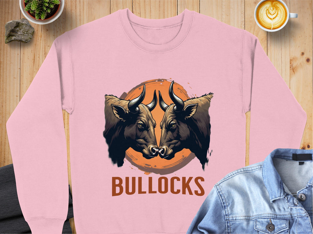 Garment Graphics presents this unisex pink sweatshirt with a striking design: two bulls facing on an orange circle, BULLOCKS printed below. Set amidst denim, a cappuccino, a plant, and a rock for artistic flair.