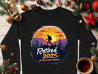A black, unisex shirt by Garment Graphics features a silhouetted hiker against a sunset and mountains, with the text Retired 2025, Not My Problem Anymore. Its displayed among Christmas decorations on wood.