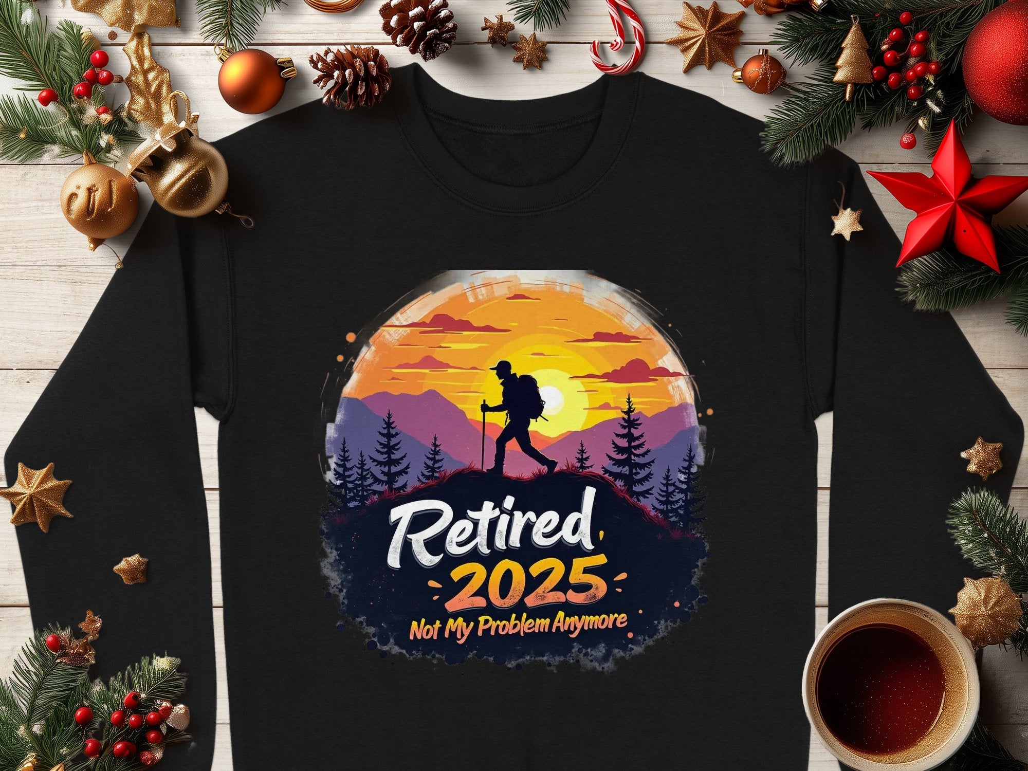 A black, unisex shirt by Garment Graphics features a silhouetted hiker against a sunset and mountains, with the text Retired 2025, Not My Problem Anymore. Its displayed among Christmas decorations on wood.