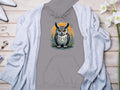 Garment Graphics gray hoodie showcases a fierce owl on an orange circular backdrop, styled with a light gray cardigan. Pair it with white pointed shoes, a gold ring, silver earrings, and white flowers on wood for a unique style. Made from medium-heavy fabric for comfort and durability.