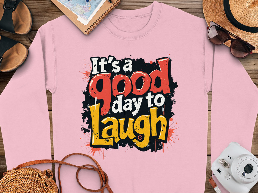 Garment Graphics pink sweatshirt features Its a good day to Laugh in bold, colorful text. Made from medium-heavy fabric, it comes in unisex sizes and is styled alongside a camera, sunglasses, straw hat, open notebook, and sandals on a wooden surface.