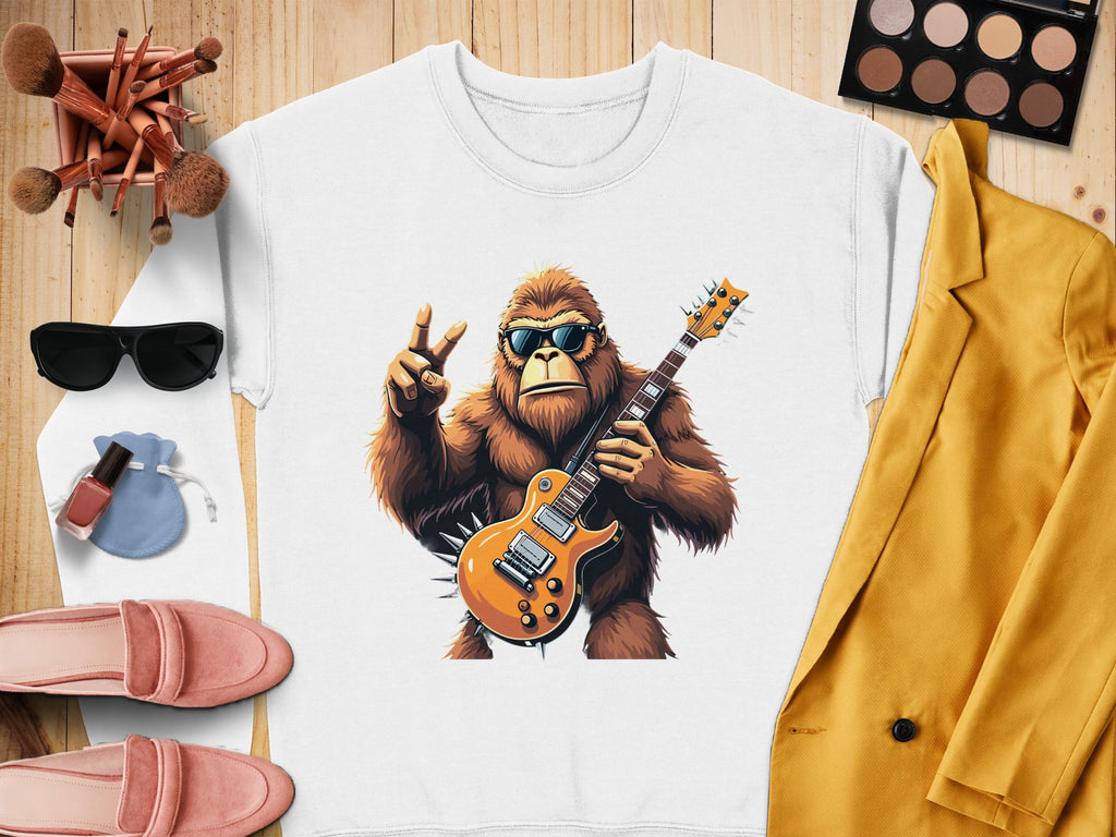 Garment Graphics sweatshirt showcases a cartoon gorilla in sunglasses playing an electric guitar, set amid makeup items, black sunglasses, pink slippers, and a yellow jacket on a wooden surface—ideal for music lovers.