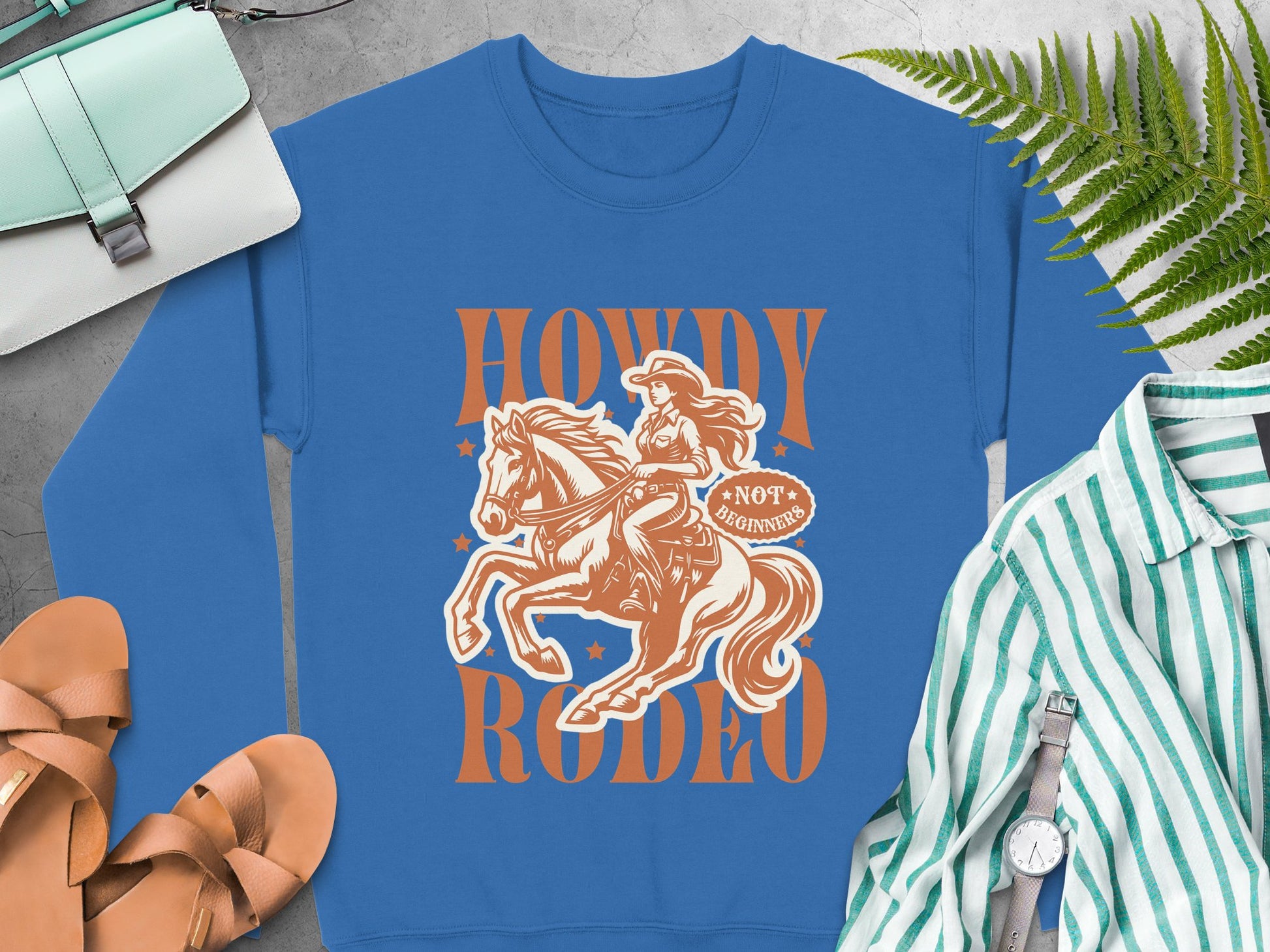 The blue sweatshirt by Garment Graphics showcases a cowboy spirit illustration with Howdy Rodeo and Not for Beginners, ideal for rodeo fans. It rests on a gray surface alongside sandals, a striped shirt, bag, watch, and ferns.