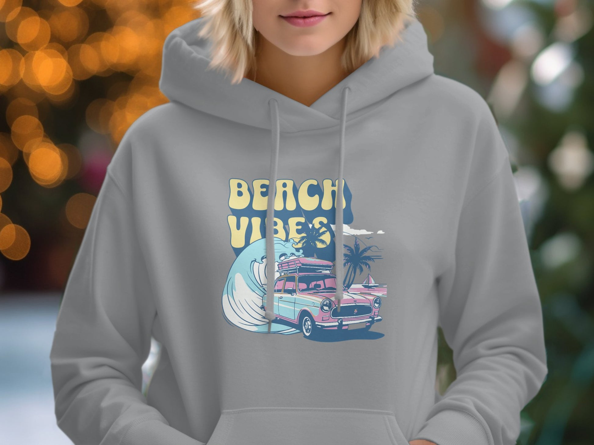 A person wears Garment Graphics gray hoodie, featuring Beach Vibes with a pink vintage car, palm trees, and a wave. The background has blurred lights and greenery, enhancing the retro beach style.