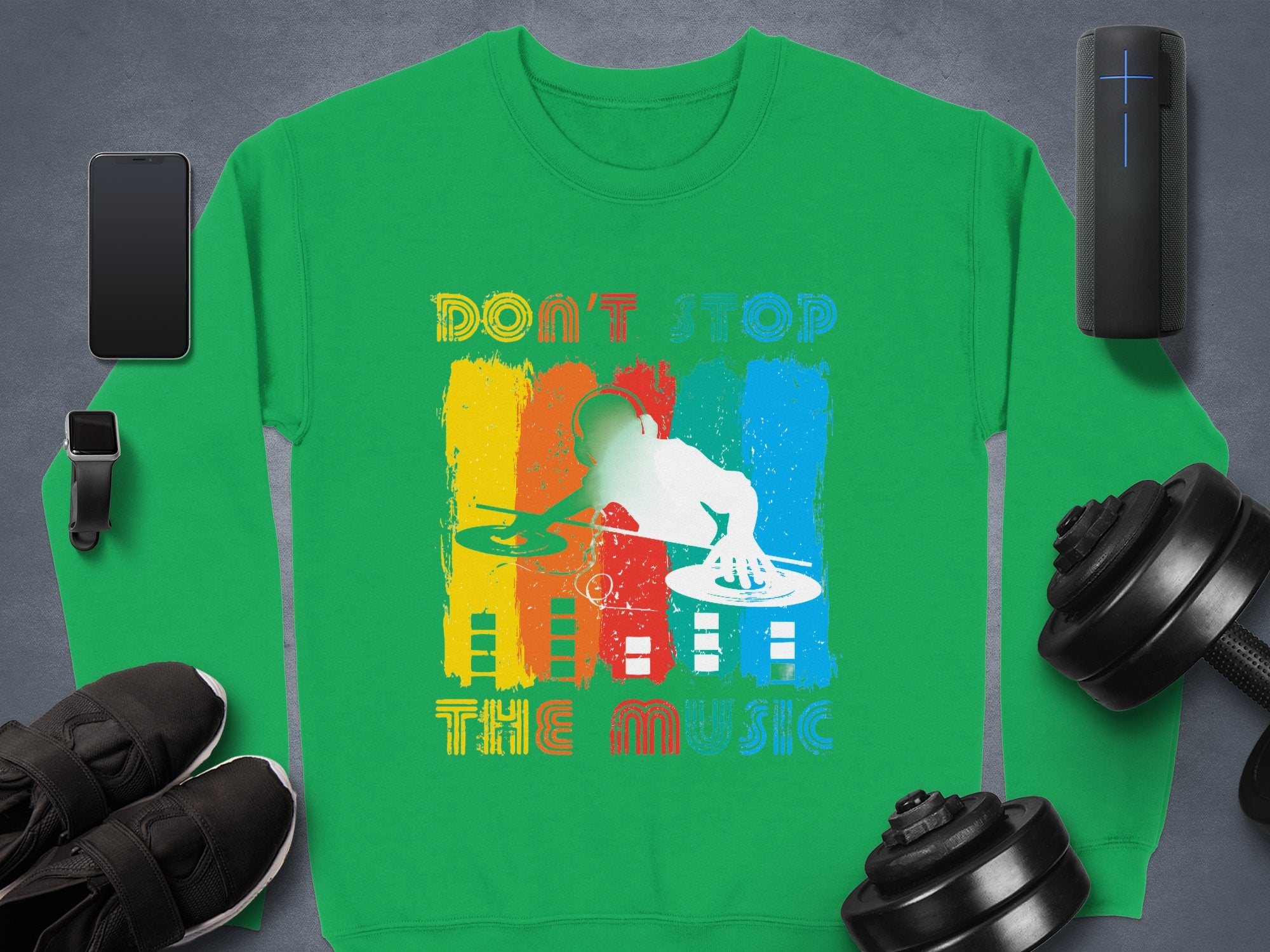 A dynamic DJ-themed green sweatshirt from Garment Graphics features Dont Stop the Music text. On a gray backdrop, it is paired with a smartphone, wireless speaker, smartwatch, sneakers, and weights—ideal for stylish music enthusiasts craving rhythm.