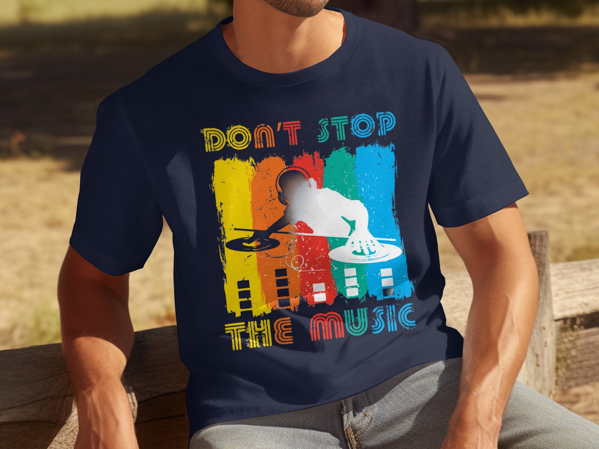 A person wears a Garment Graphics navy T-shirt with a DJ silhouette and colorful stripes. The text reads Dont Stop the Music. They sit on an outdoor wooden bench, ideal for music lovers.