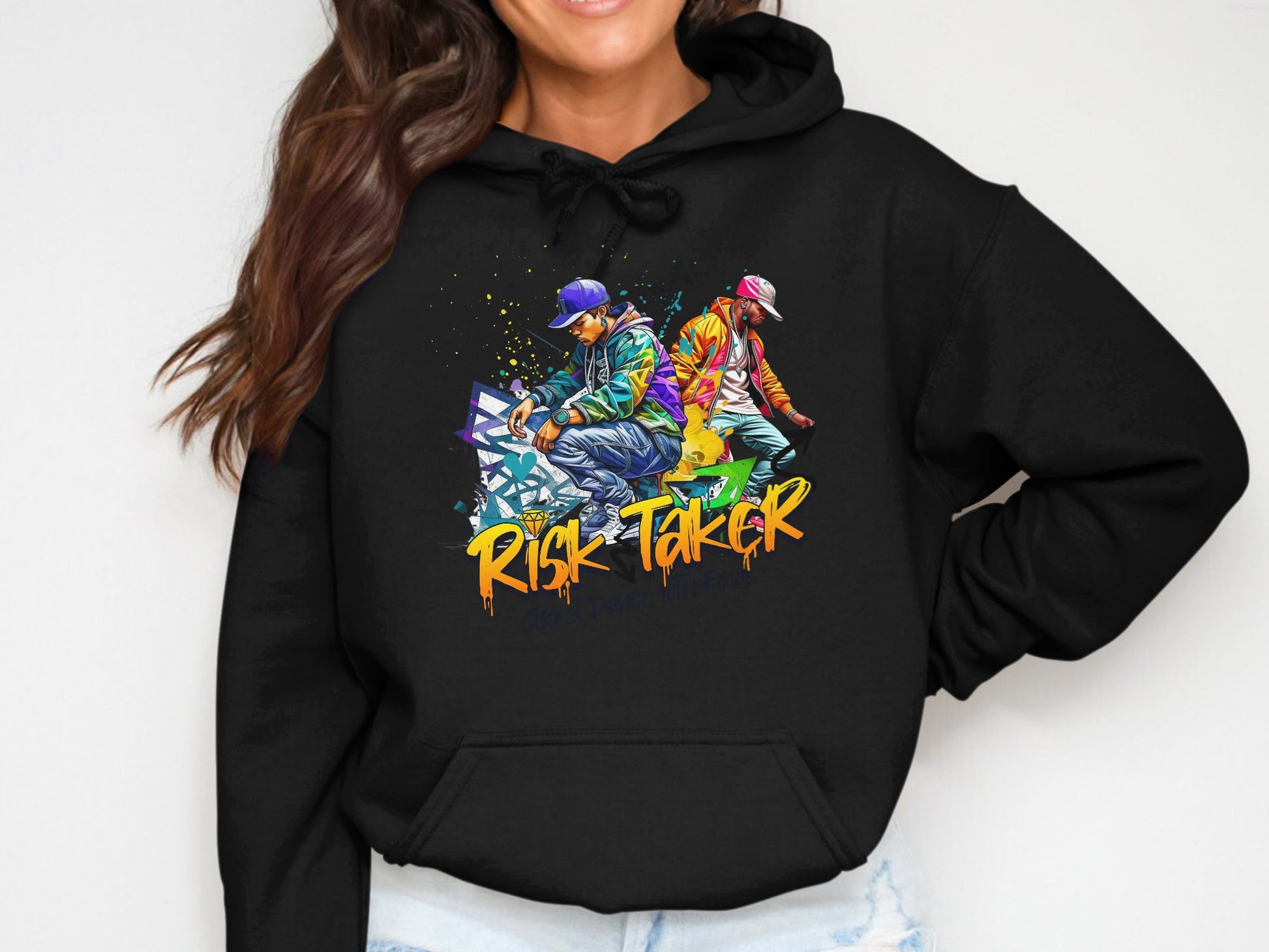 The individual is wearing a black graphic hoodie by Garment Graphics featuring vibrant art of three people in urban streetwear. Boldly stylish, the piece displays Risk Taker Street Dance beneath. They smile, with one hand casually in a pocket.