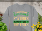 A gray sweatshirt from Garment Graphics, showcasing a bold and humorous design, reads If an Englishman gets run down by a truck, he apologizes to the truck in lively green, yellow, and black letters. Unisex sizing offers a perfect fit for everyone. Its surrounded by green plants and yellow flowers.