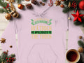 A light-colored hoodie by Garment Graphics lies on a wooden surface among Christmas ornaments and coffee, featuring a humorous quote about apologizing to a truck that ran you down, highlighting English manners with wit.