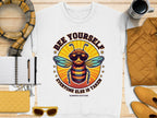 A white unisex Garment Graphics T-shirt with a bee in sunglasses and Bee Yourself Everyone Else is Taken text pairs perfectly with essentials like a notebook, scarf, and yellow quilted jacket for embracing your unique style.