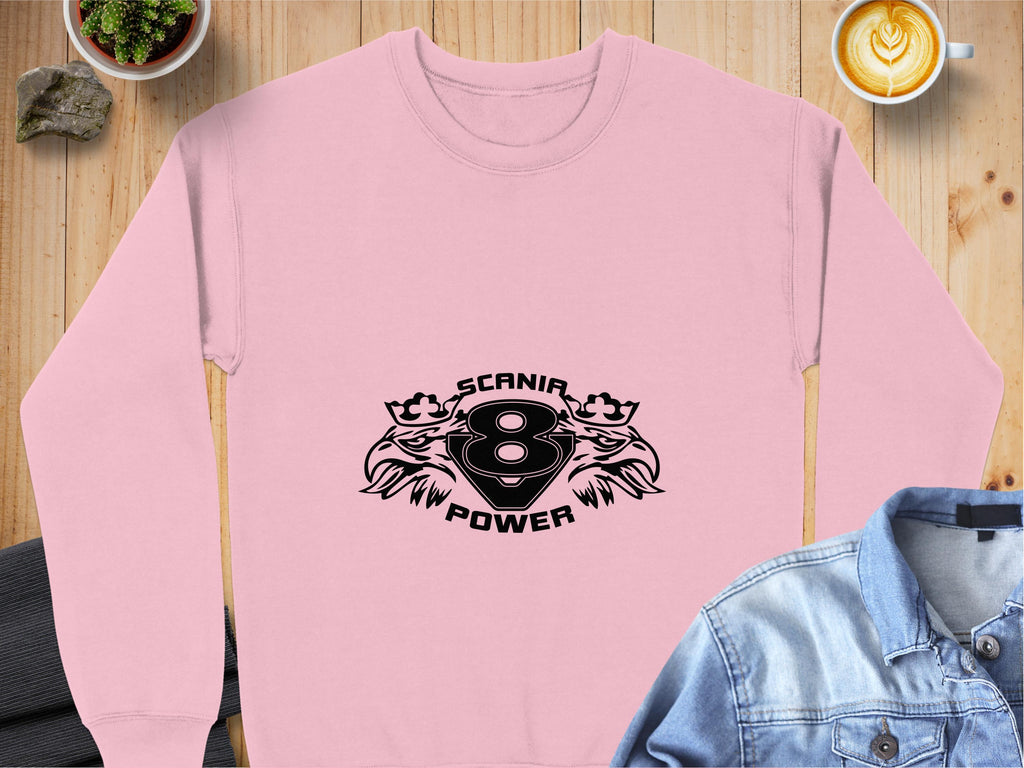 A Garment Graphics pink sweatshirt with the Scania Power 8 logo and two stylized animal heads lays on a wooden surface. Nearby are a cappuccino, denim jacket, suit jacket, small plant, and rock.