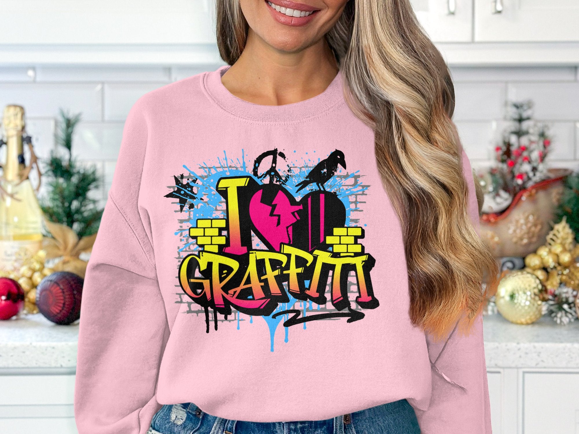 Wearing a Garment Graphics artistic sweatshirt, a person sports a pink top with an urban graffiti design that says I Love Graffiti, featuring bricks, splash effects, and paint drips, standing in a kitchen adorned with holiday decor.