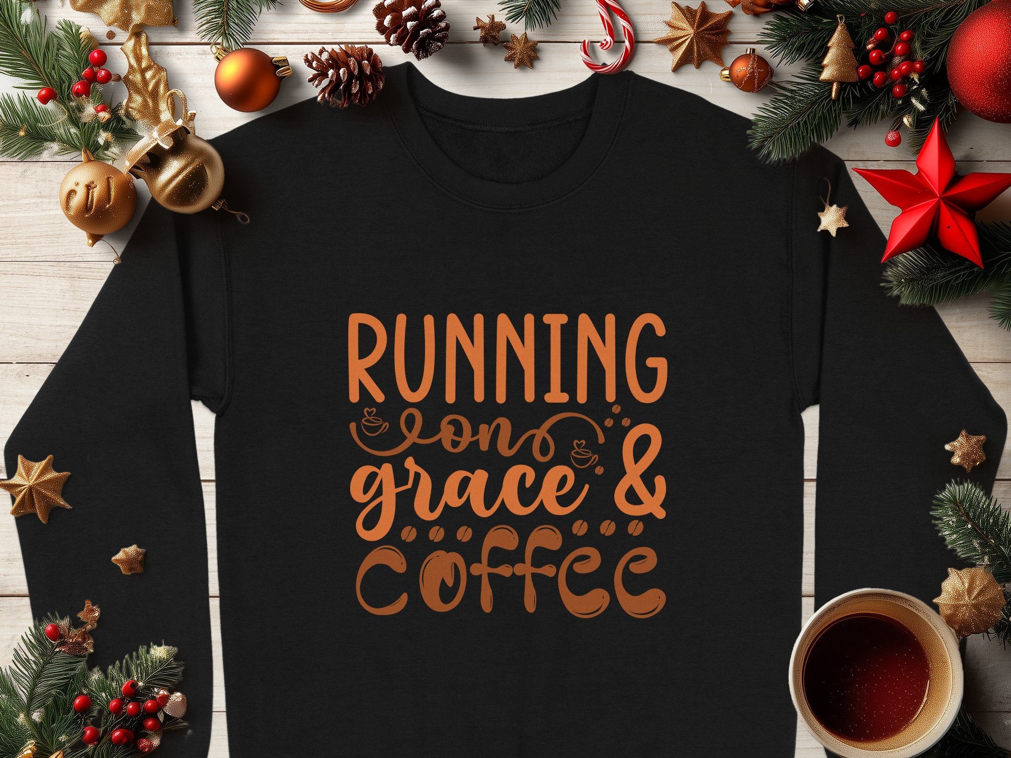 Garment Graphics black graphic sweatshirt features Running on grace & coffee in brown text with cup illustrations, surrounded by pine branches, ornaments, and a coffee cup on white wood. It exudes cozy vibes and is available in unisex sizes.