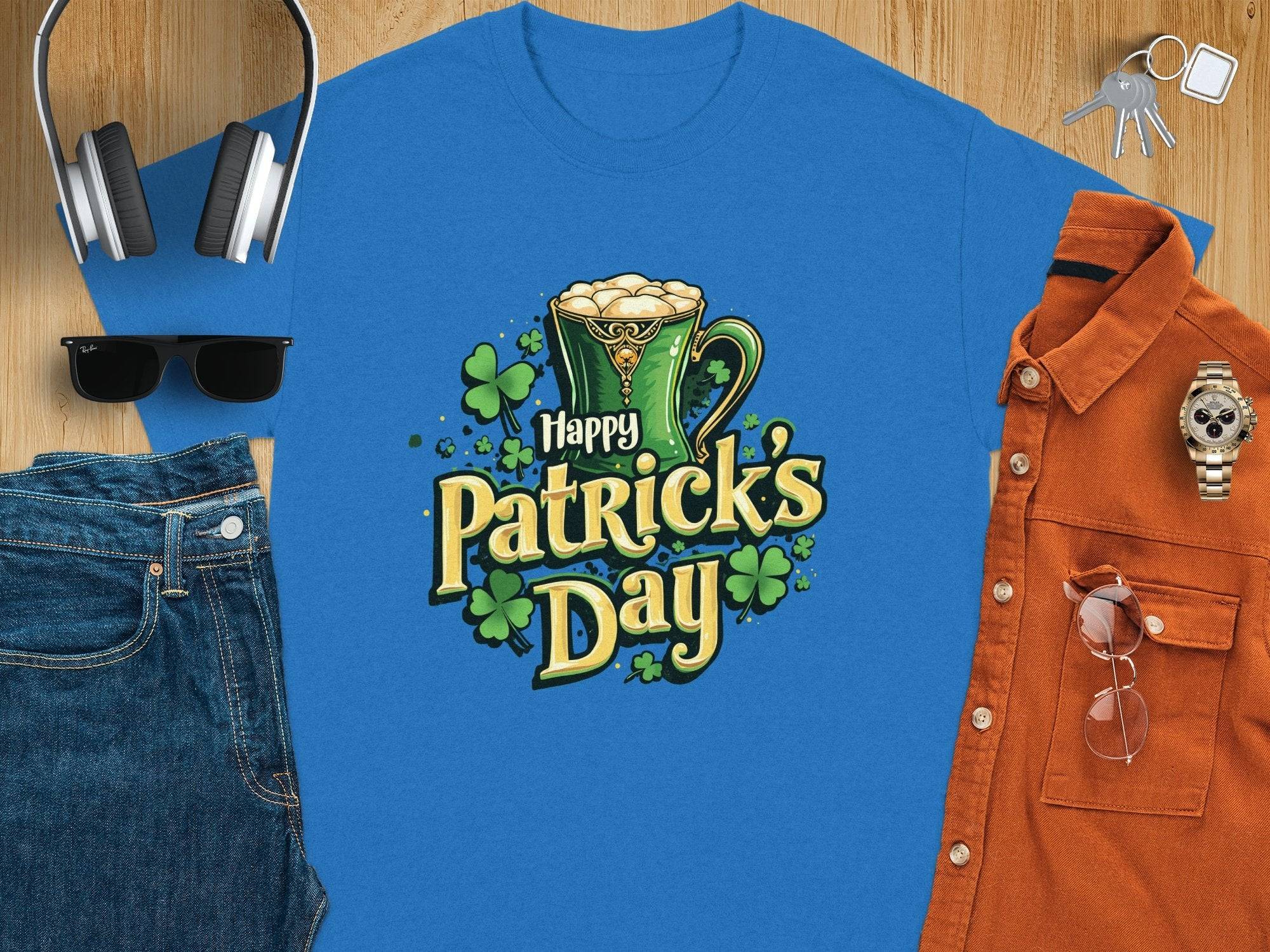 A festive flat lay by Garment Graphics features a blue Happy Patricks Day T-shirt with a green mug, surrounded by sunglasses, headphones, denim jeans, an orange shirt, wristwatch, smartphone, and keys on the wooden surface—a stylish and celebratory scene.