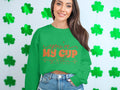 A woman wearing a cozy Garment Graphics sweatshirt with Youre My Cup of Tea in orange stands against a shamrock-adorned wall. Her long, dark hair flows over her jeans. The unisex-sized sweatshirt perfectly complements the festive vibe.