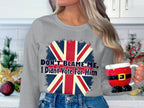 A person with long hair holds a Santas pants mug, wearing a Garment Graphics gray unisex sweatshirt featuring the Union Jack and Dont Blame Me, I Didnt Vote For Him text. Festive decorations adorn the kitchen counter in the background.