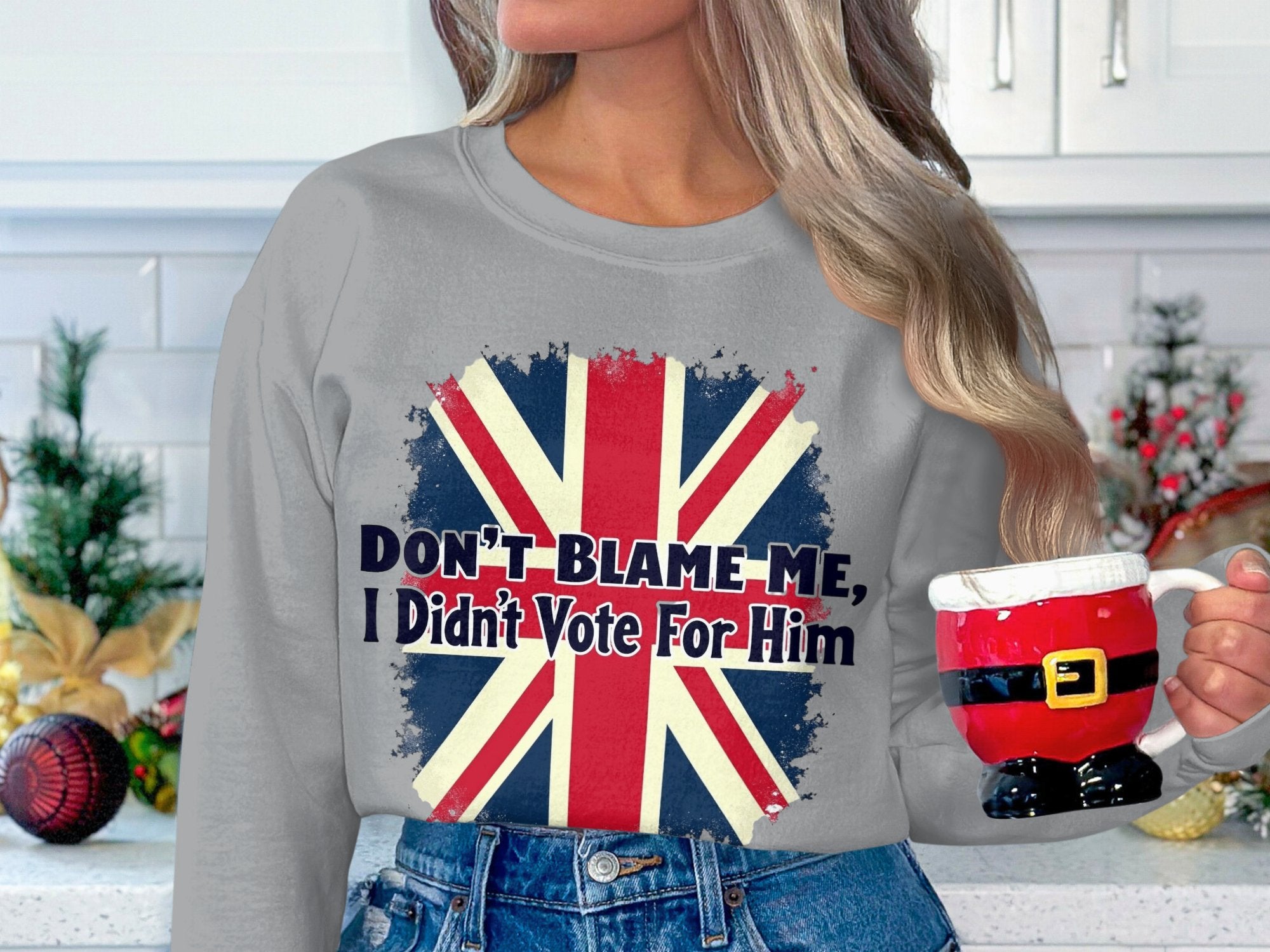 A person with long hair holds a Santas pants mug, wearing a Garment Graphics gray unisex sweatshirt featuring the Union Jack and Dont Blame Me, I Didnt Vote For Him text. Festive decorations adorn the kitchen counter in the background.