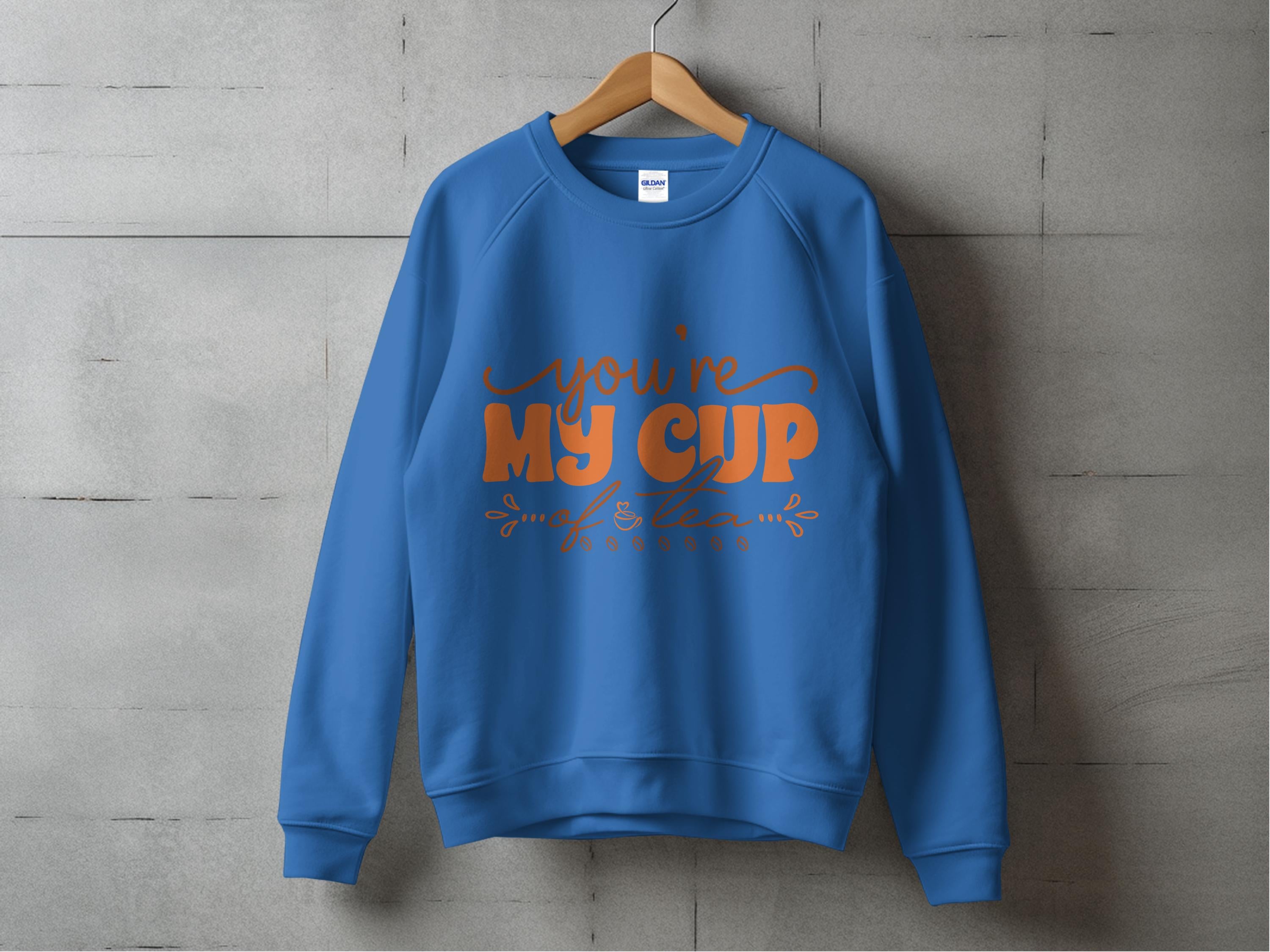 A cozy unisex sweatshirt by Garment Graphics hangs on a wooden hanger against a concrete wall, featuring an elegant orange Youre My Cup of Tea design in cursive.