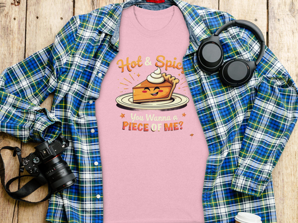 The Garment Graphics pink t-shirt with a hot and spicy pie design—featuring a pie with a face, arms, whipped cream, saying Hot & Spicy and You Wanna a Piece of Me?—is placed on wood alongside a blue plaid shirt, headphones, camera, and coffee cup.