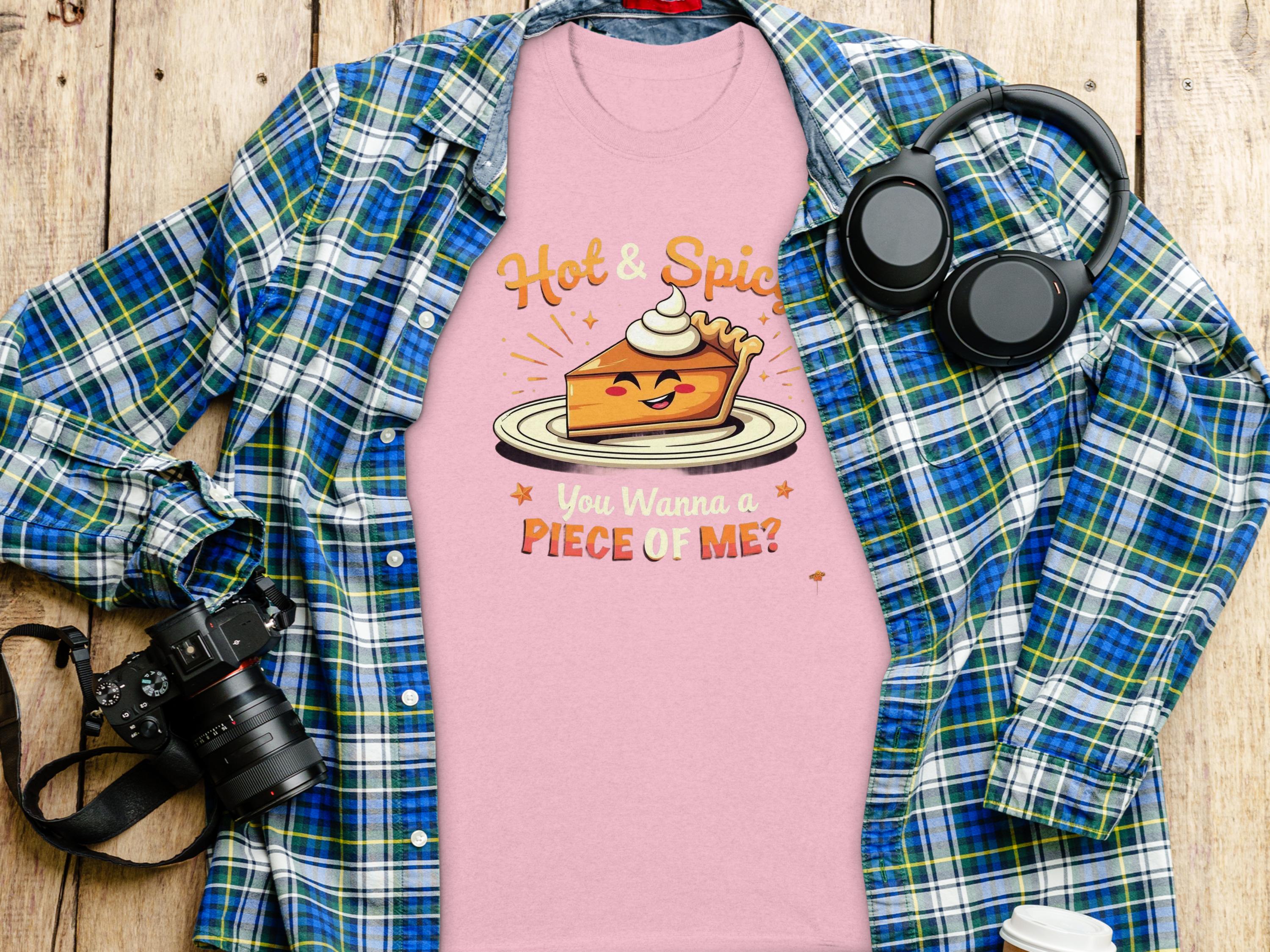 The Garment Graphics pink t-shirt with a hot and spicy pie design—featuring a pie with a face, arms, whipped cream, saying Hot & Spicy and You Wanna a Piece of Me?—is placed on wood alongside a blue plaid shirt, headphones, camera, and coffee cup.