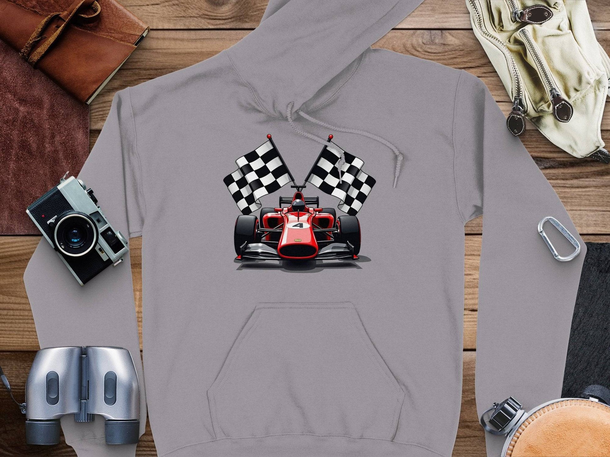 A Garment Graphics gray hoodie with a racing car design, bold red and checkered flags is displayed on wood. Nearby are a camera, binoculars, leather-bound book, adventurous bag with papers, and a carabiner.