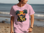 A beachgoer dons a Garment Graphics pink t-shirt featuring palm trees, a tropical sunset, and ONE LOVE text. The tranquil ocean stretches behind them, creating an idyllic scene for beach enthusiasts.