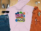 A laid-out outfit includes a Garment Graphics pink Lets Make Some Noise hoodie, making a bold statement, paired with blue jeans, sunglasses, headphones, a burnt orange shirt, watch, and keys on a wooden surface.