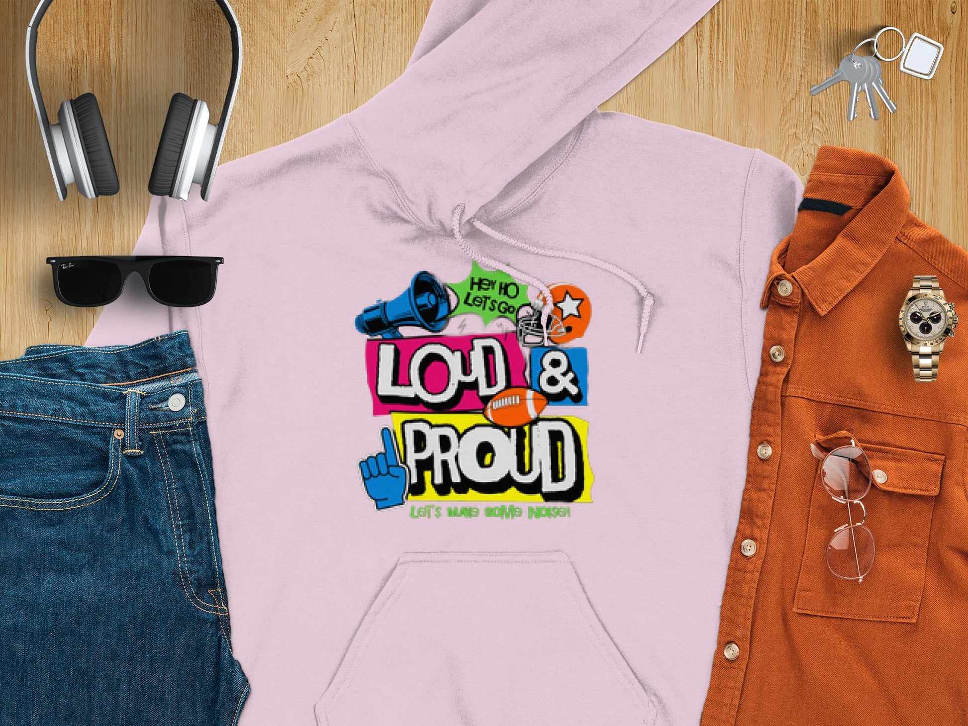 A laid-out outfit includes a Garment Graphics pink Lets Make Some Noise hoodie, making a bold statement, paired with blue jeans, sunglasses, headphones, a burnt orange shirt, watch, and keys on a wooden surface.