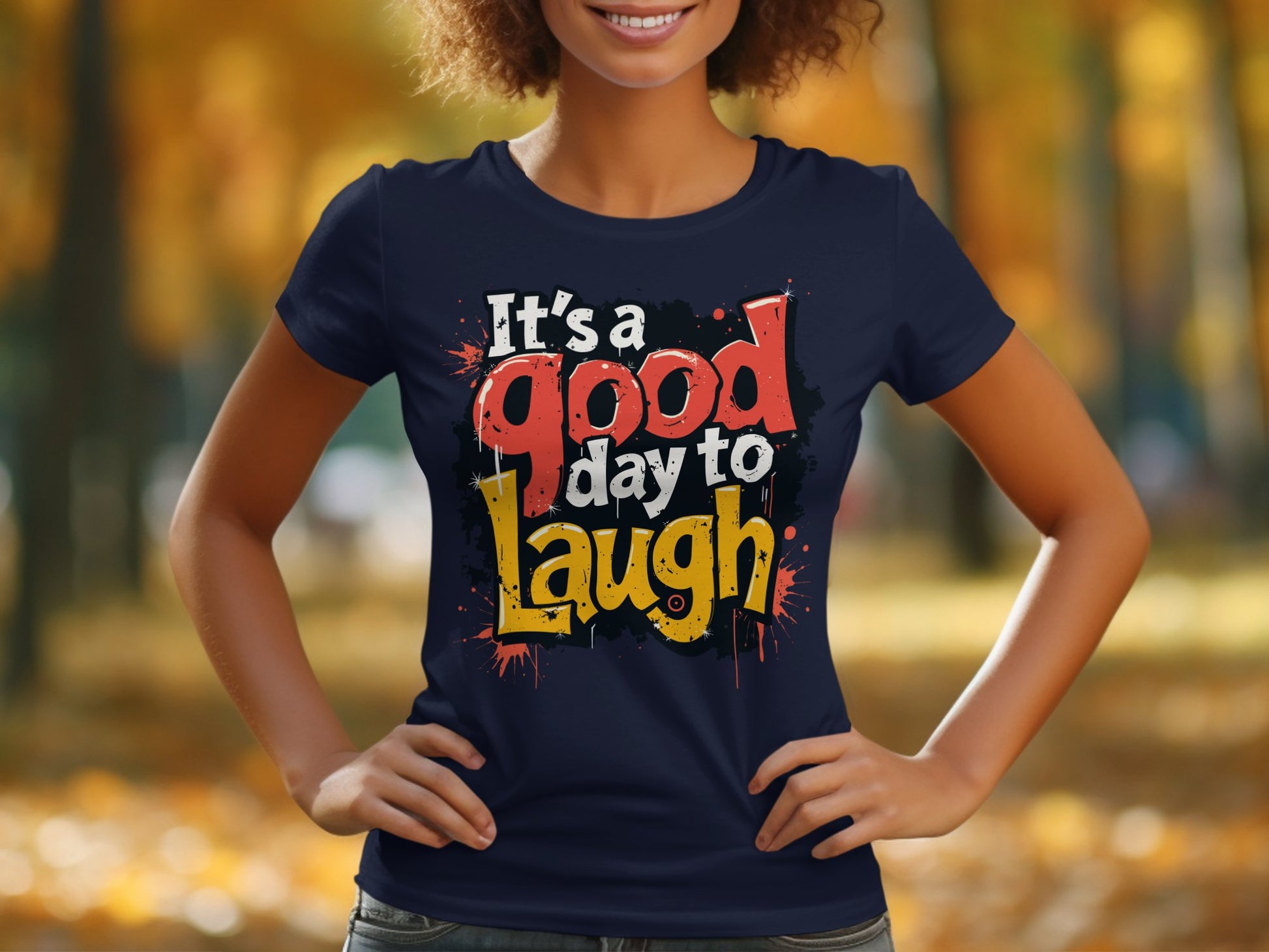 A person outdoors wears a Garment Graphics semi-fitted navy blue T-shirt with a vibrant Its a good day to laugh print, surrounded by blurred autumn trees and sunlight. The scene exudes positivity as they smile with hands on hips.