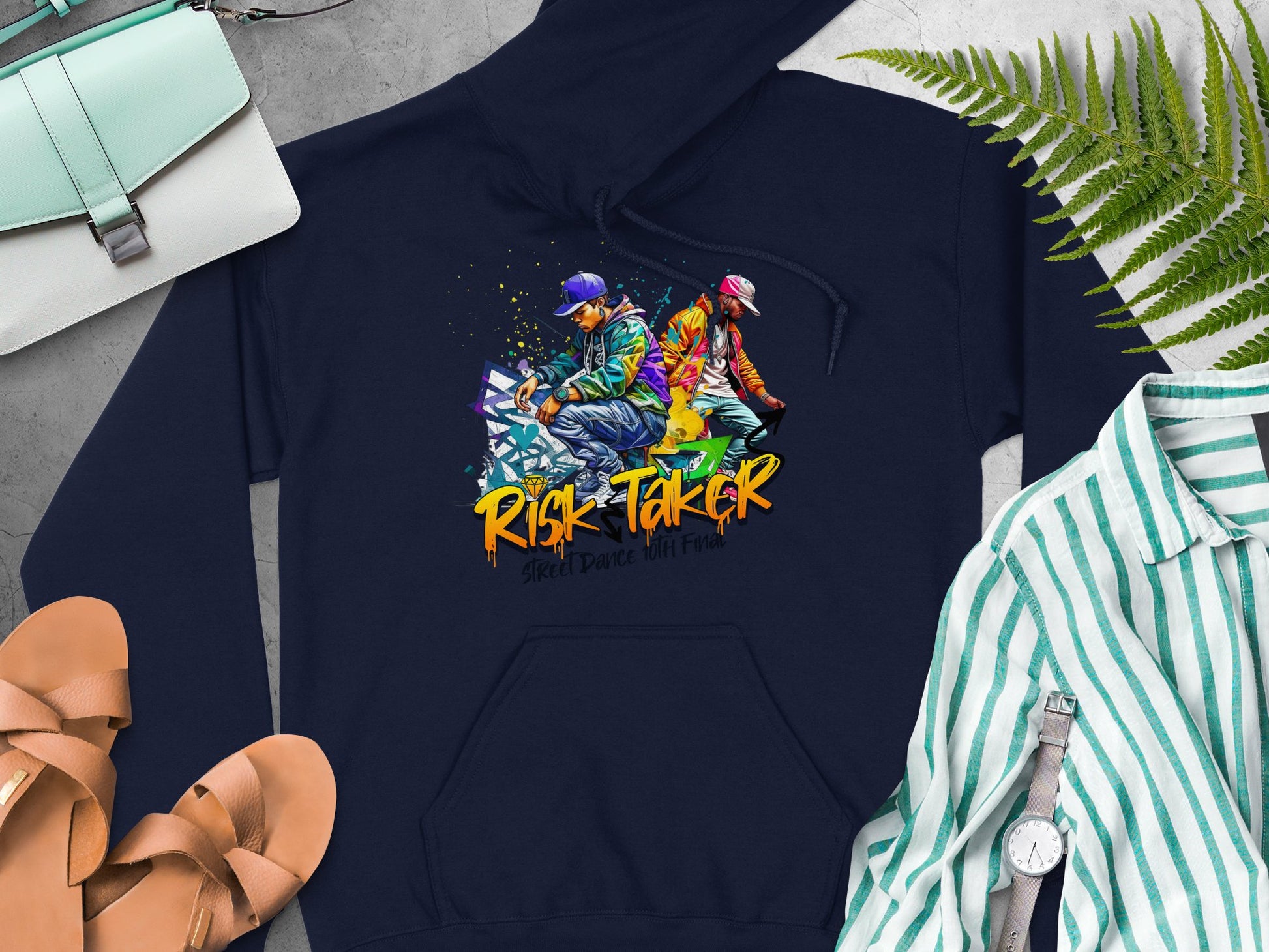 The Garment Graphics navy blue graphic hoodie features two dancers with Risk Taker and Street Dance with Fire. This bold look is completed with a green bag, tan sandals, a striped shirt, a watch, and fern leaves.