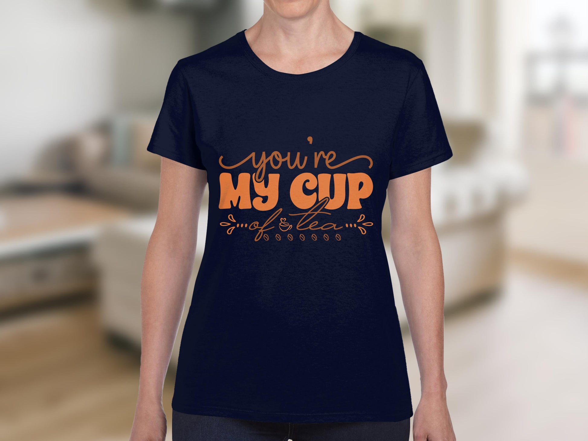 A person in a dark blue Garment Graphics T-shirt made from 100% cotton, with vibrant orange text saying Youre My Cup of Tea, stands indoors against a softly blurred living room.