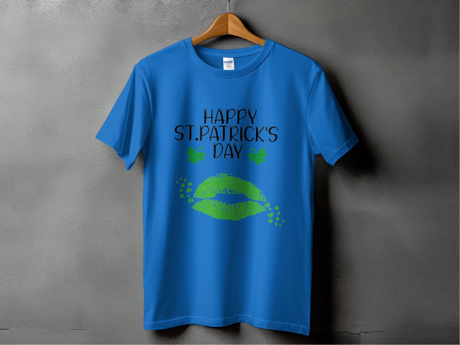 A Garment Graphics T-shirt on a wooden hanger features bold green lips, two clovers, and the festive Happy St. Patricks Day text against a gray background.