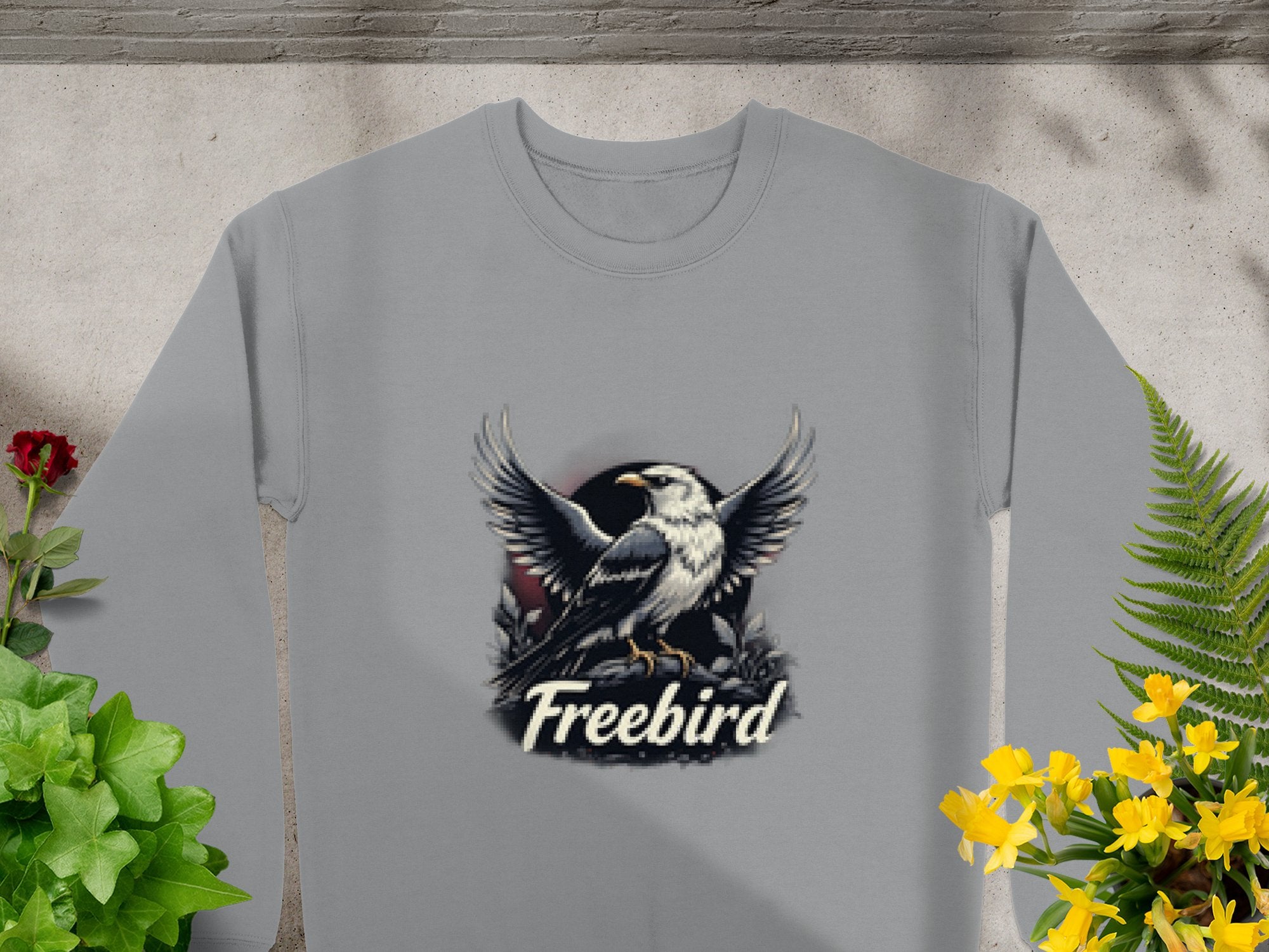 A gray sweatshirt by Garment Graphics showcases an eagle with outstretched wings above Freebird. Surrounded by green and yellow plants and a red flower, this unisex Freebird Graphic Sweatshirt offers casual comfort for everyone.