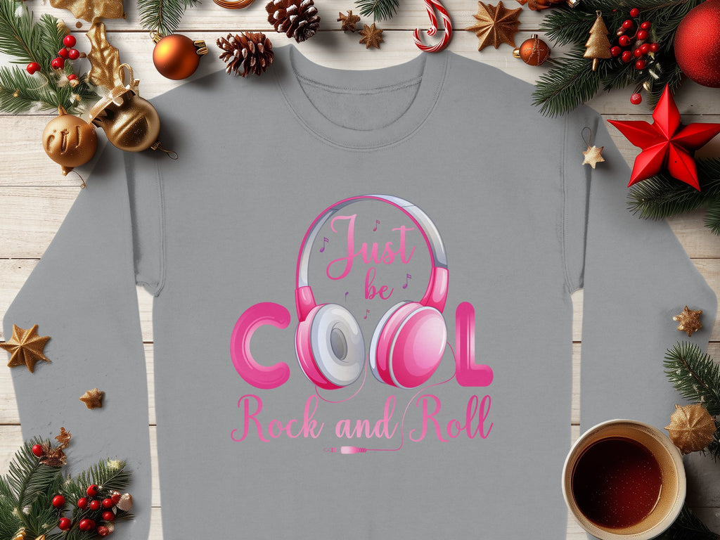 A Garment Graphics gray sweatshirt featuring Rock and Roll Pink Headphones and Just be Cool Rock and Roll lies on a wooden surface with Christmas baubles, pine branches, a red star, and coffee—a stylish choice for music lovers.