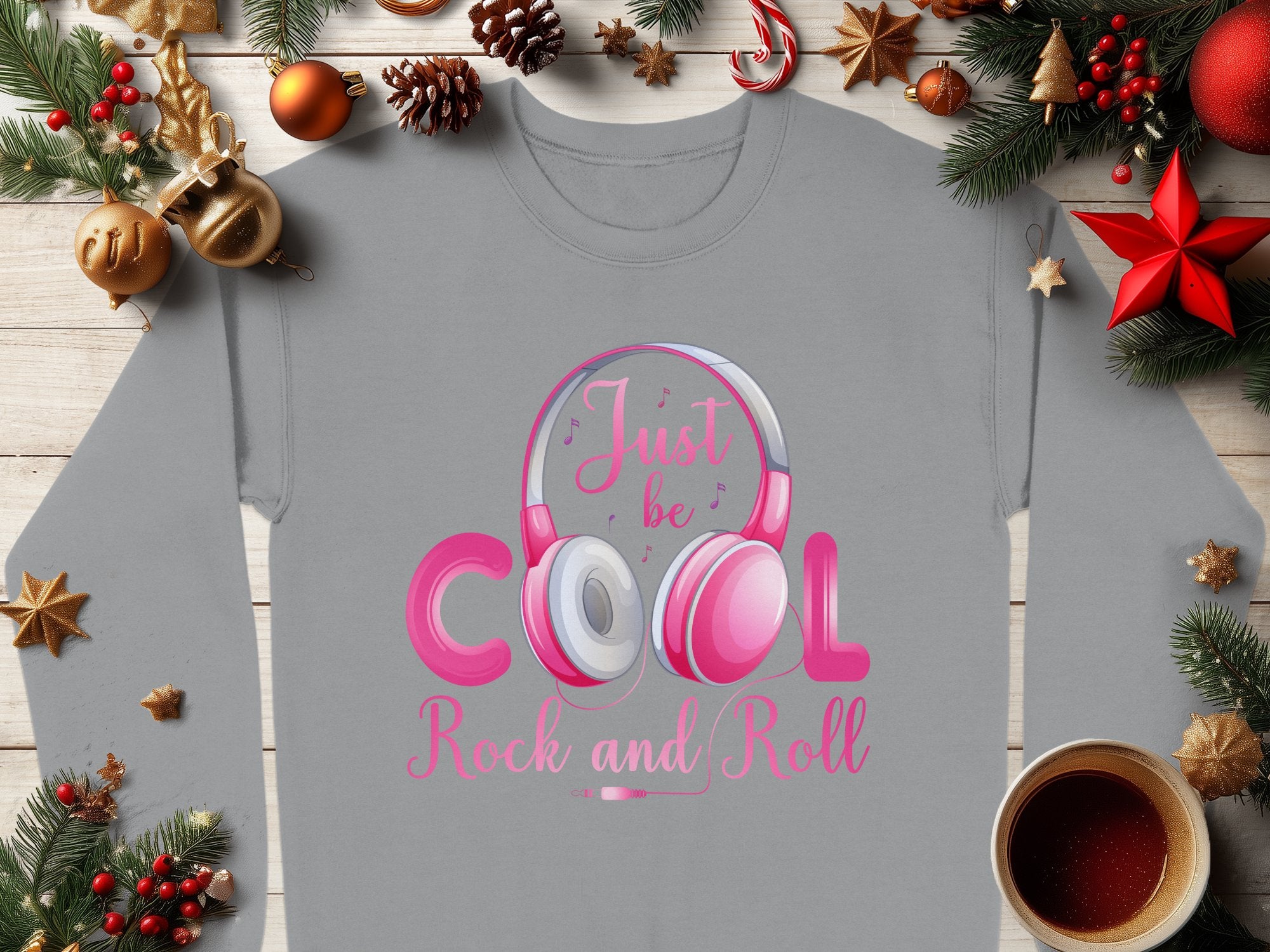 A Garment Graphics gray sweatshirt featuring Rock and Roll Pink Headphones and Just be Cool Rock and Roll lies on a wooden surface with Christmas baubles, pine branches, a red star, and coffee—a stylish choice for music lovers.