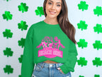 A woman with long hair wears a green Garment Graphics sweatshirt featuring pink silhouettes and the words Always Party Beach Girl, standing before a white wall decorated with green shamrocks.