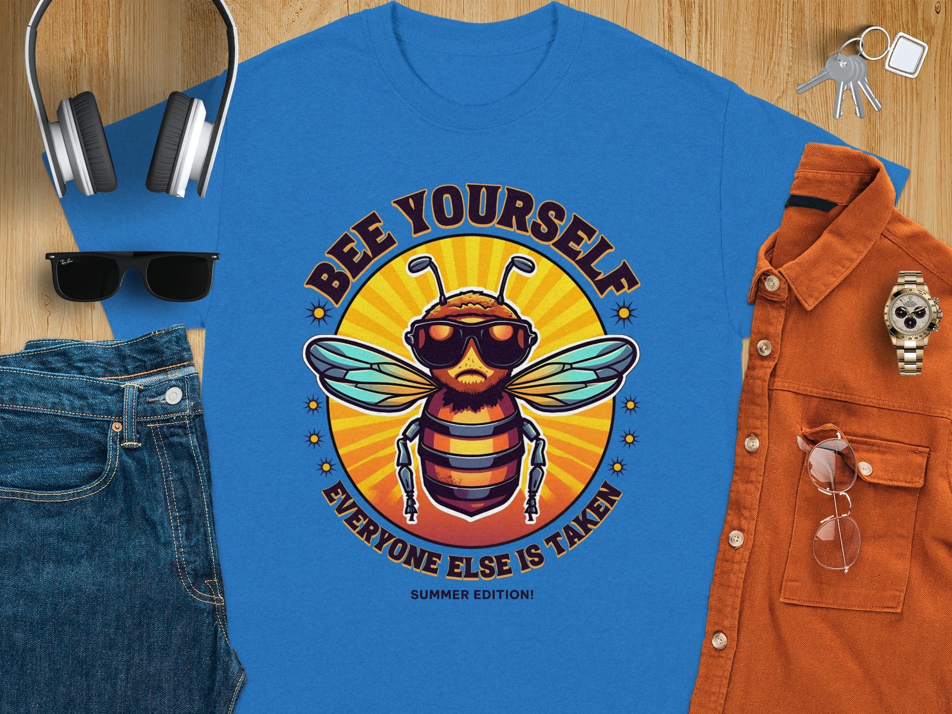 Garment Graphics offers a blue t-shirt with a bee in sunglasses and a hat, displaying Bee Yourself, Everyone Else Is Taken, Summer Edition! Available in family sizes. Styled with jeans, sunglasses, headphones, keys, an orange shirt, and a watch on wooden background.