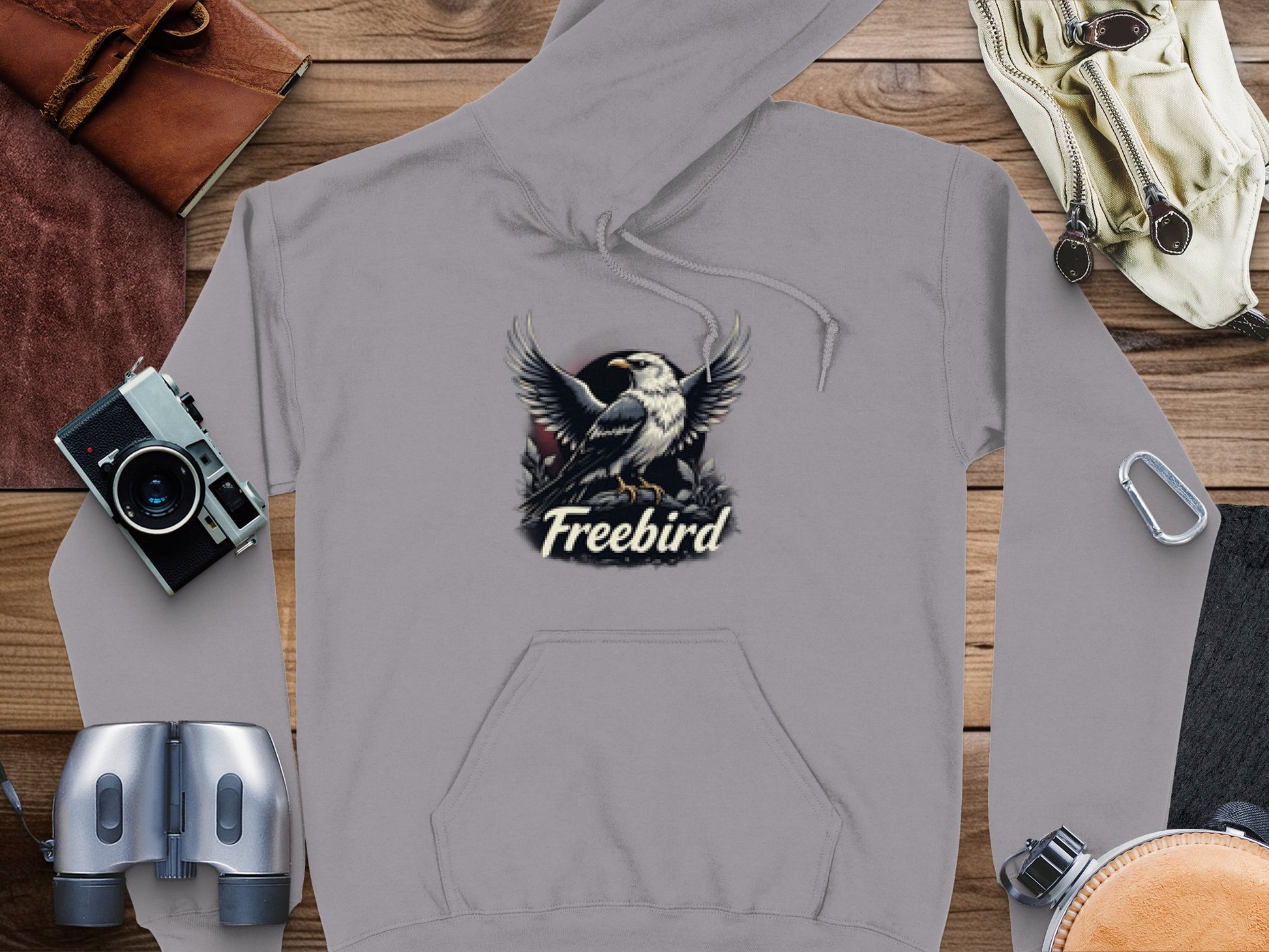 Garment Graphics Gray Freebird Graphic Pullover Hoodie features an eagle with outstretched wings above Freebird, surrounded by items like a camera, binoculars, hat, and gloves on a wooden surface for a distinctive look.