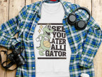 The Garment Graphics white t-shirt showcases a playful alligator with a basketball and the words See You Later Alligator. Its arranged on a blue/green plaid shirt with a camera, headphones, and drink cup on wood—ideal for those who appreciate unique graphic apparel.