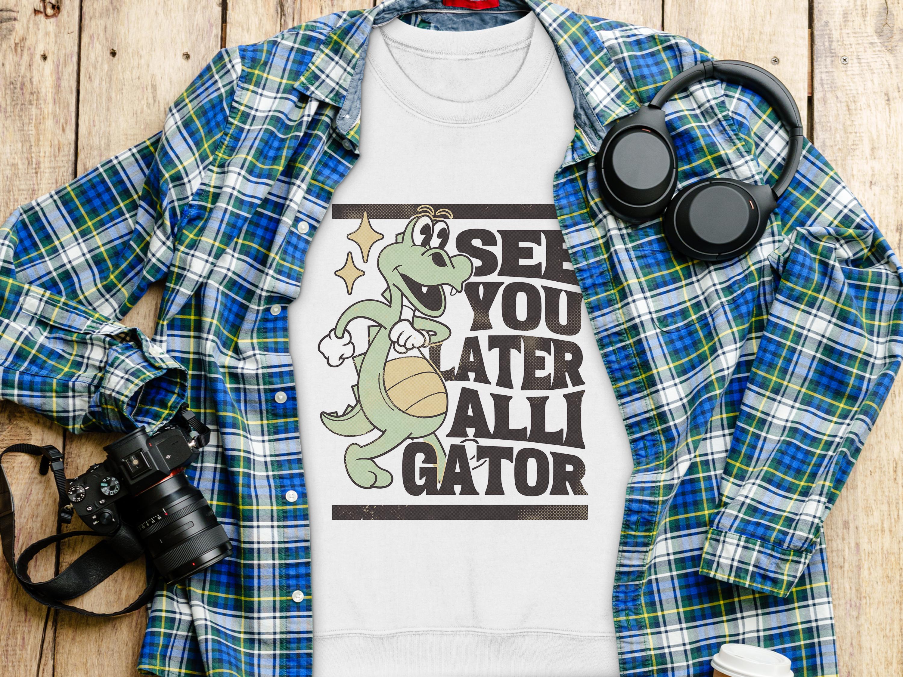 The Garment Graphics white t-shirt showcases a playful alligator with a basketball and the words See You Later Alligator. Its arranged on a blue/green plaid shirt with a camera, headphones, and drink cup on wood—ideal for those who appreciate unique graphic apparel.